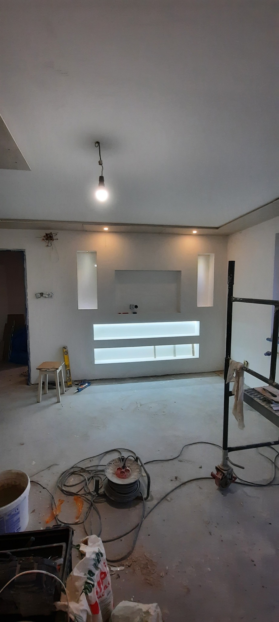 Wnętrze w trakcie wykańczania: ściany przygotowane pod gładzie, oświetlenie LED wnękowe, widoczne narzędzia i materiały budowlane, surowe wykończenie podłogi i stropu.