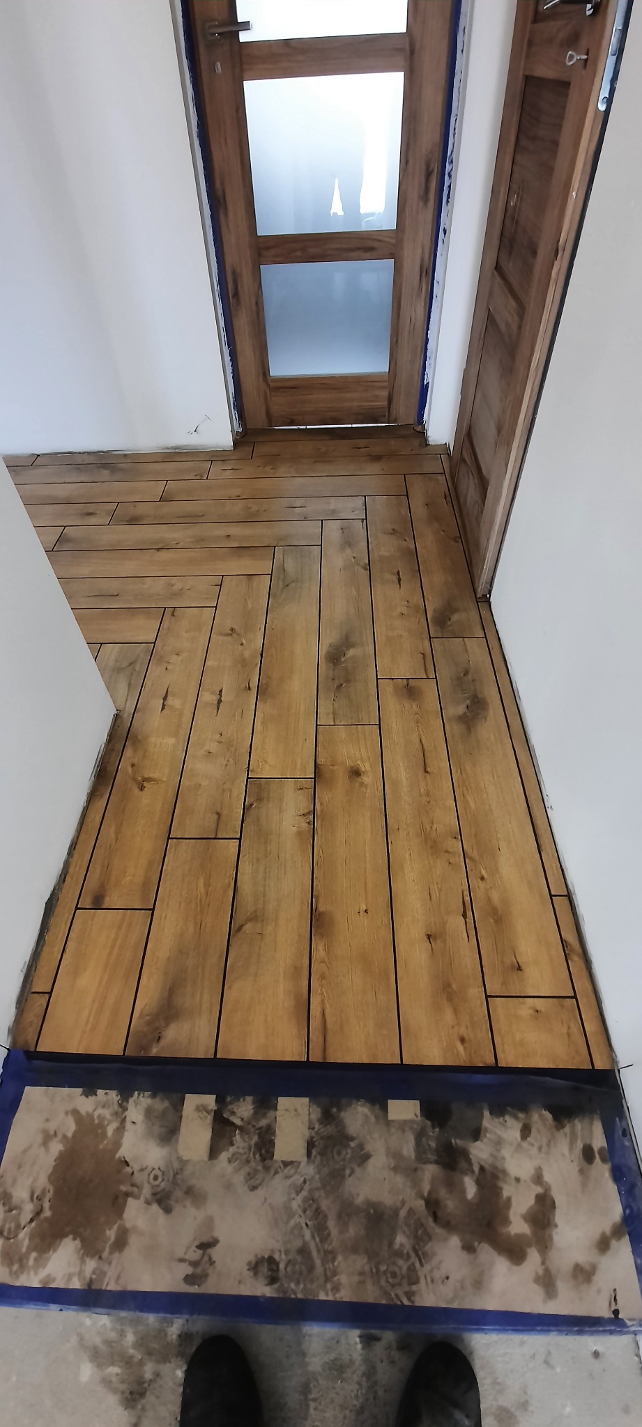 Ułożona podłoga z paneli imitujących drewno w jodełkę, widok z góry na fragment podłogi przy drzwiach z szybami, zabezpieczona folią malarską przy krawędziach, w tle widoczne stopy fotografa.