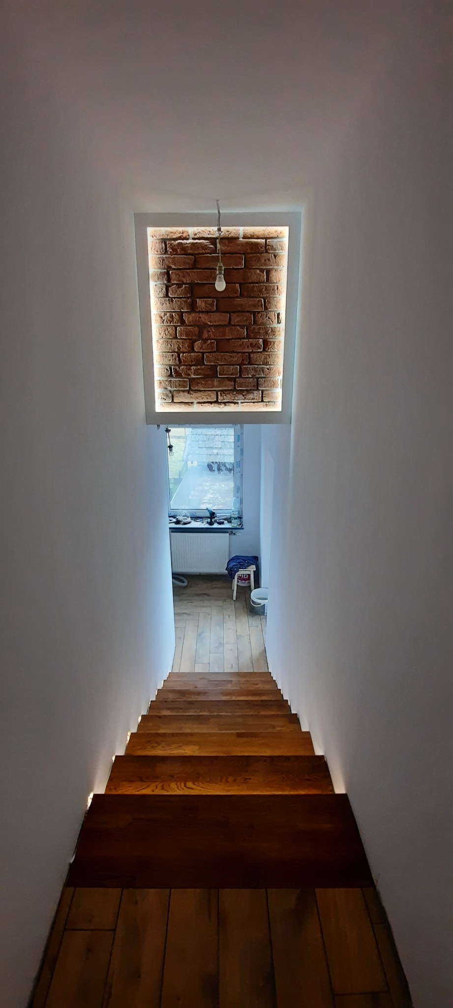 Wnętrze klatki schodowej z drewnianymi schodami, oświetleniem LED pod stopniami i dekoracyjną ścianą z cegły podświetloną lampą wiszącą.