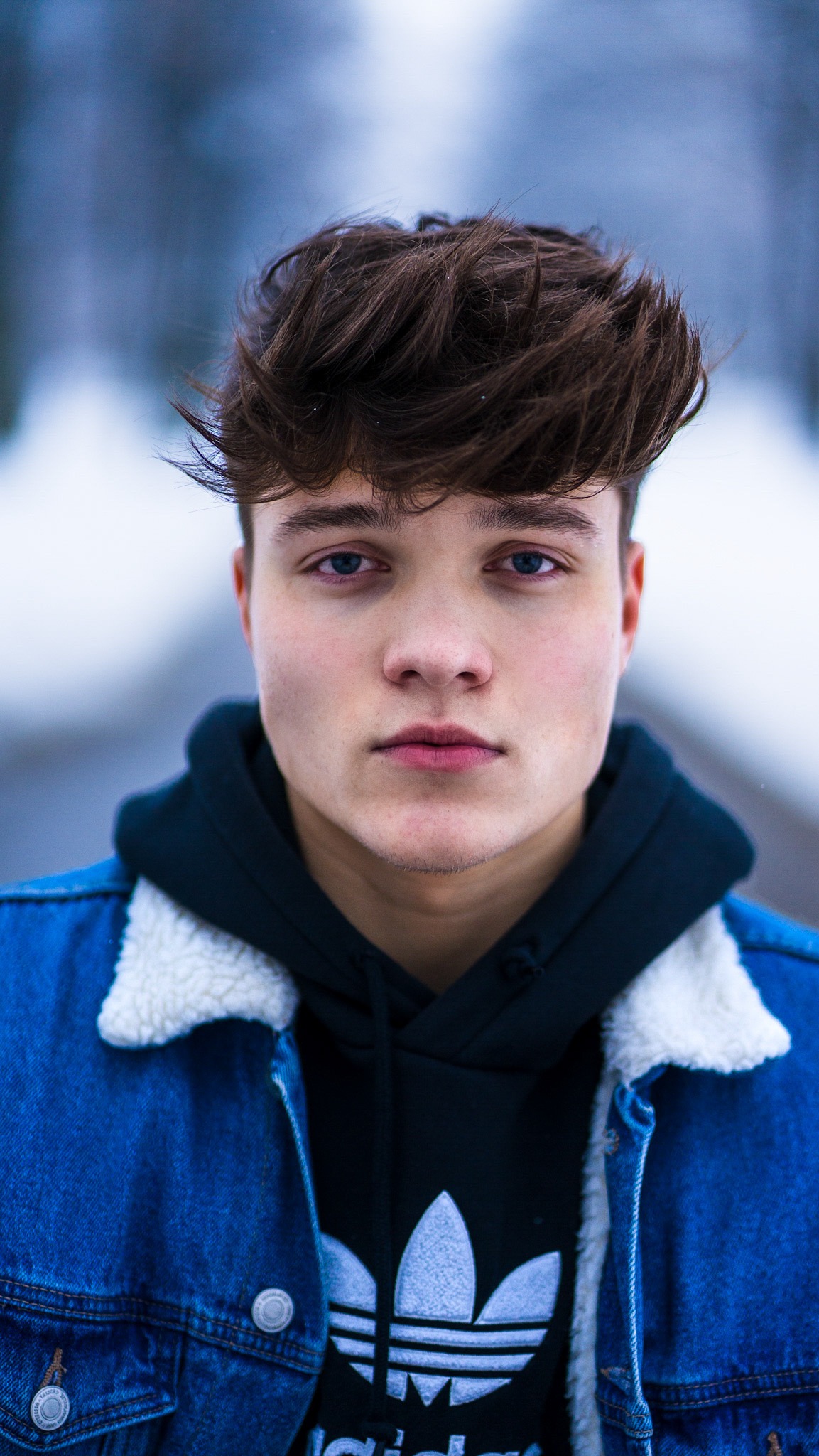 Portret młodego mężczyzny w Radzionkowie, ubrany w dżinsową kurtkę i czarną bluzę z kapturem z logo Adidas, na rozmytym tle zimowego krajobrazu.