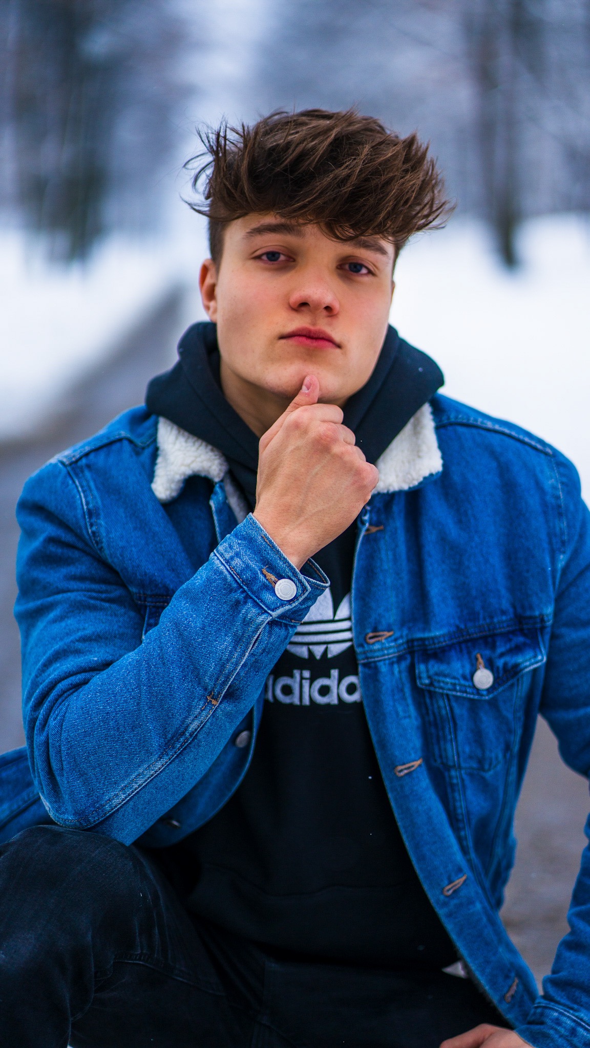 Młody mężczyzna w dżinsowej kurtce i czarnej bluzie z kapturem, pozujący na tle zimowego krajobrazu, z dłonią pod brodą.