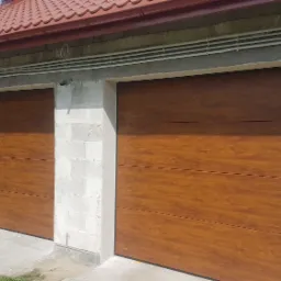 Dwie brązowe bramy garażowe segmentowe o strukturze drewna, zamontowane w surowym budynku z betonu komórkowego, widoczne przewody elektryczne pod dachem.