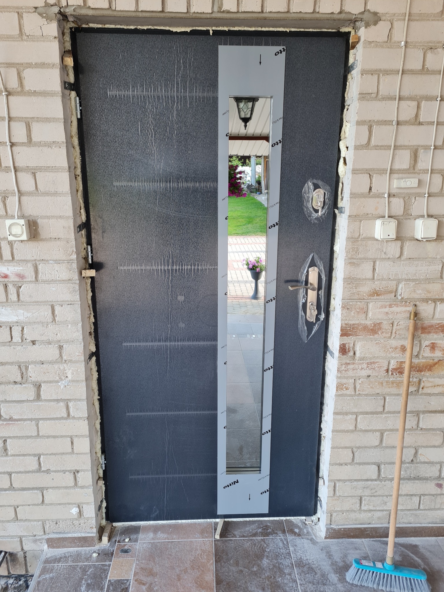 Montaż zewnętrznych drzwi wejściowych z podłużnym przeszkleniem; widoczne uszczelnienia pianką montażową i folia ochronna na klamce, w tle mur z cegły i ogród.