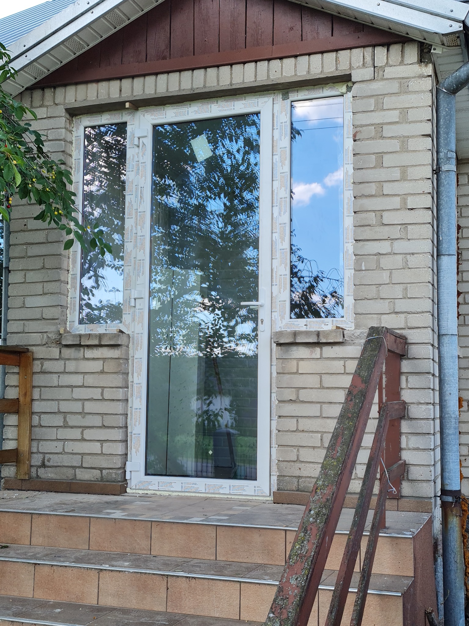 Zestaw okien i drzwi balkonowych z białymi ramami, zamontowany w elewacji z jasnej cegły, widok z zewnątrz na schody wejściowe.