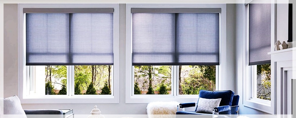 Nowoczesne okno z plisowanymi osłonami w kolorze szarym, widok na zielony ogród, niebieski fotel w salonie.