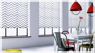 Jasne wnętrze jadalni z trzema oknami osłoniętymi roletami w zygzakowaty wzór, czerwone lampy wiszące nad białym stołem z różnokolorowymi krzesłami.