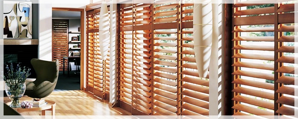 Jasne wnętrze z drewnianymi żaluzjami poziomymi, widoczne przez okno drzewa, w tle nowoczesny fotel i obraz nad kominkiem.