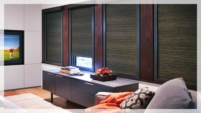 Nowoczesny salon z trzema oknami wyposażonymi w poziome żaluzje w ciemnych odcieniach brązu, zamontowane w drewnianych ramach, widoczne zza sofy i szafki RTV.