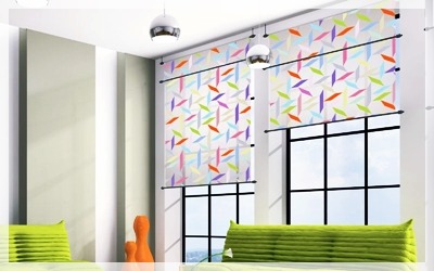 Nowoczesne okno z dwoma panelami materiału w geometryczne wzory, zawieszonymi na czarnych rurkach, w jasnym wnętrzu z zielonymi poduszkami.