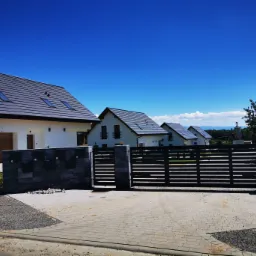 Nowoczesne, parterowe domy z szarymi dachami i czarnymi ramami okiennymi, widoczne zza ogrodzenia z kamiennymi słupkami i drewnianymi przęsłami, na tle błękitnego nieba.