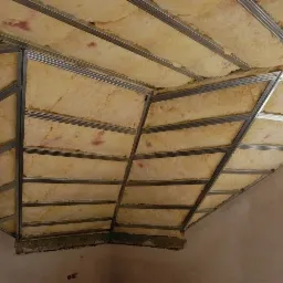 Skośny sufit poddasza z izolacją z wełny mineralnej zamontowaną w metalowym stelażu, widoczne niedokończone ściany i okno.