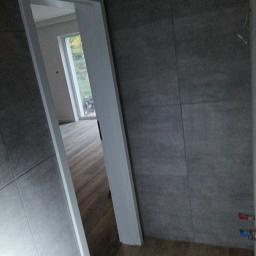 Probud - Wnętrze łazienki w trakcie wykańczania: szare płytki na ścianach, drewnopodobne panele na podłodze, biała ościeżnica drzwiowa z widokiem na dalszą część mieszkania z parkietem i oknem.