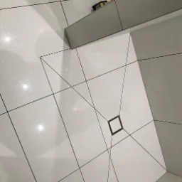 Białe płytki podłogowe ułożone w geometryczny wzór z czarnymi fugami, centralnie umieszczony odpływ liniowy w kształcie kwadratu, widok z góry.