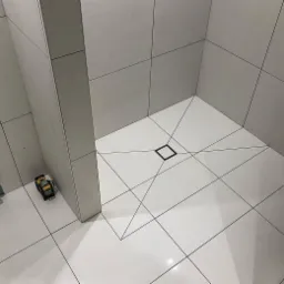 Biała, kwadratowa kabina prysznicowa z odpływem liniowym w kształcie kwadratu, wyłożona jasnymi płytkami z ciemną fugą, widoczny laser krzyżowy do poziomowania.