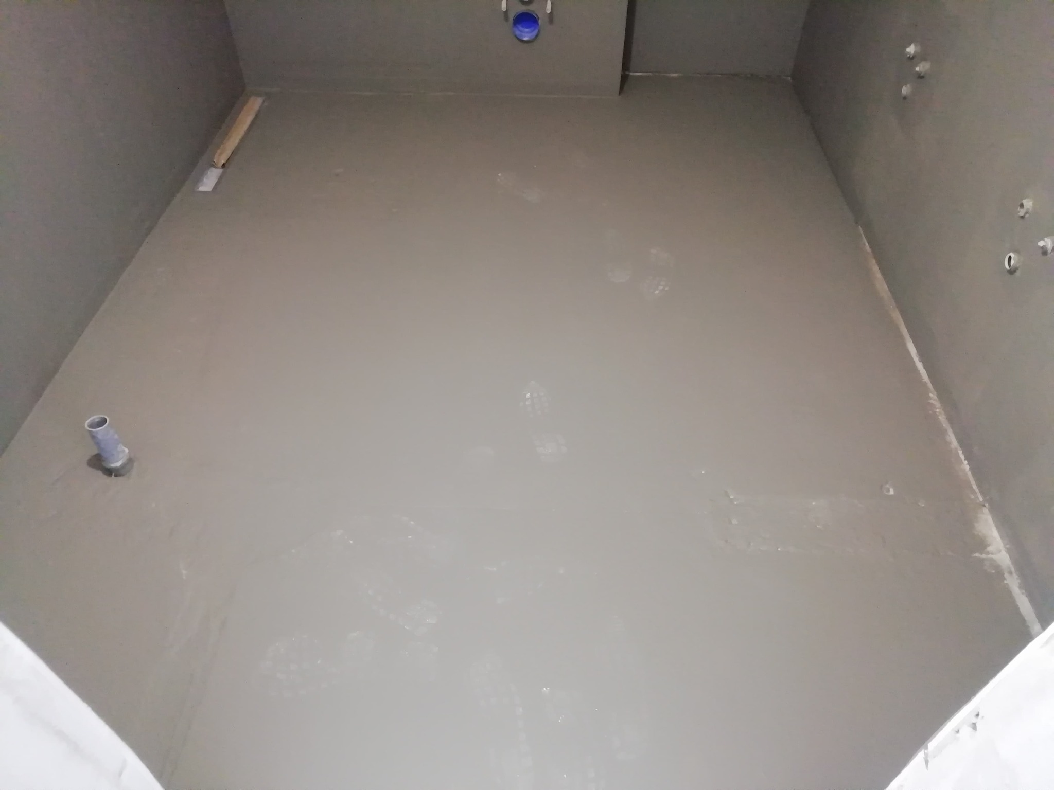 Świeżo wylana, szara wylewka samopoziomująca w łazience, widoczne ślady stóp, rura odpływowa w rogu, przygotowane otwory w ścianie na instalacje, listwa przypodłogowa w rogu.