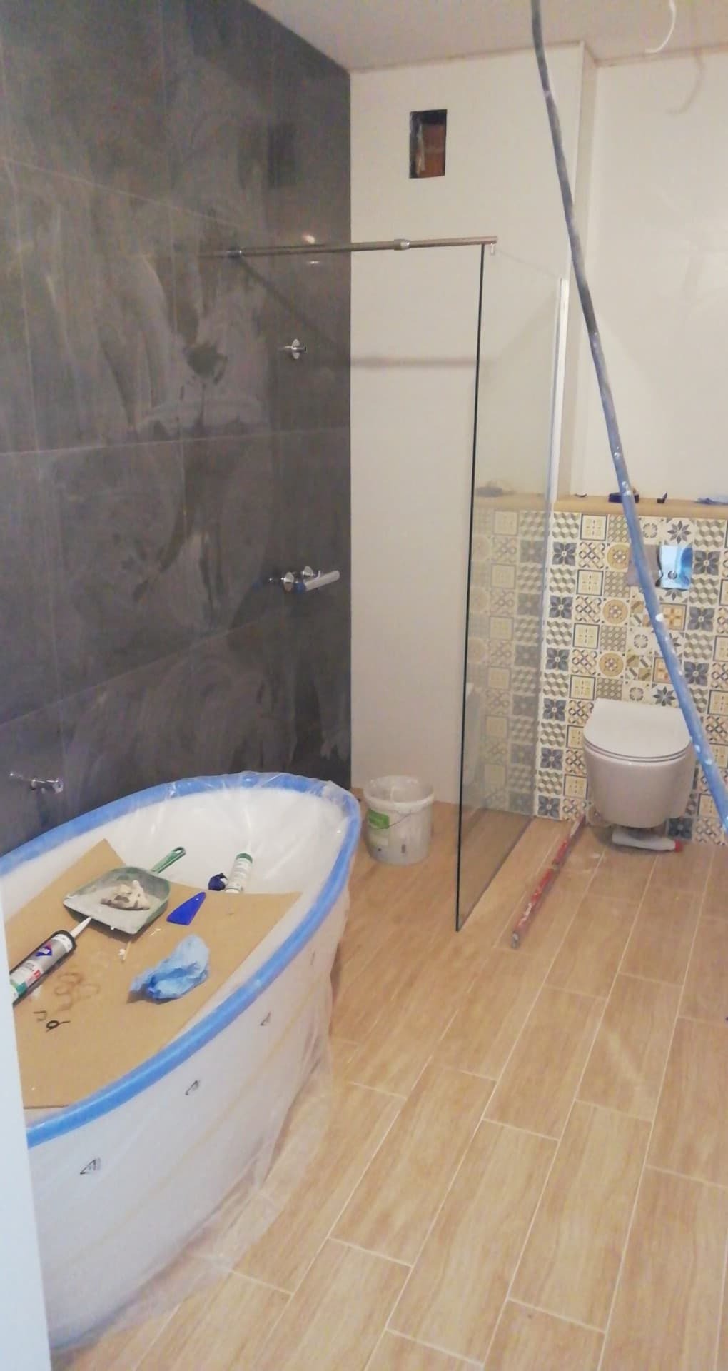 Łazienka w trakcie prac wykończeniowych: obudowana wanna zabezpieczona folią, szklana kabina prysznicowa zamontowana, widoczne narzędzia i materiały budowlane, płytki na ścianach w dwóch różnych...