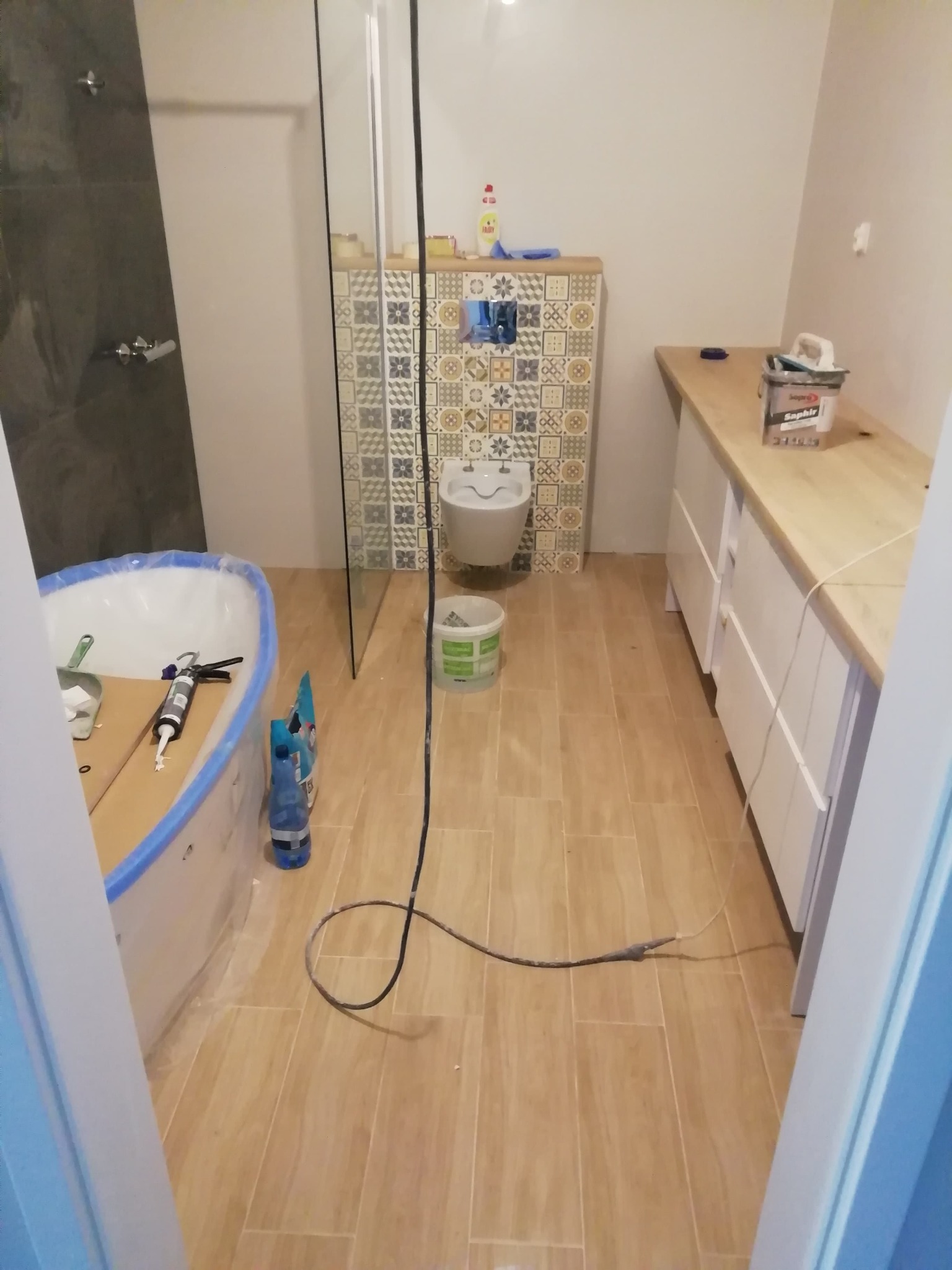 Łazienka w trakcie wykończenia: wanna z zabezpieczającą folią, częściowo ułożone płytki dekoracyjne za toaletą, drewnopodobne płytki na podłodze, białe szafki pod umywalkę z blatem z jasnego drewna...