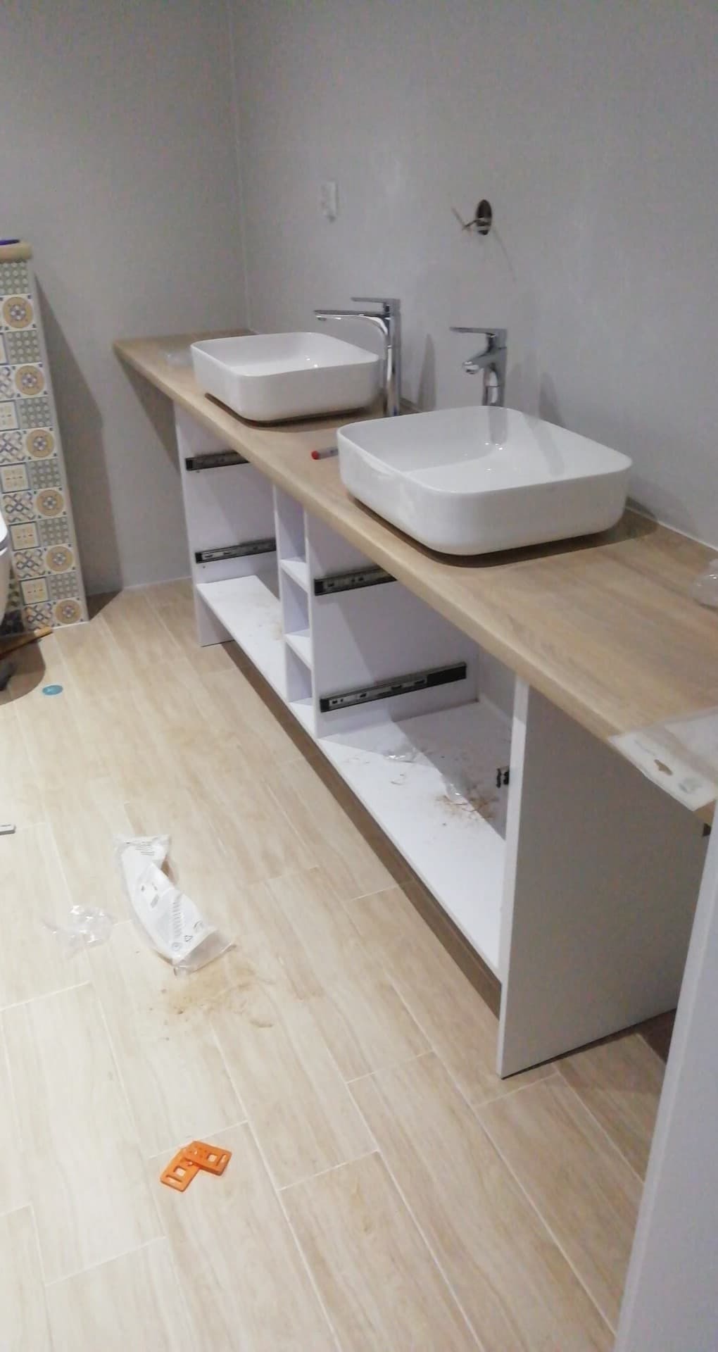 Dwie kwadratowe umywalki nablatowe z chromowanymi bateriami zamontowane na drewnianym blacie z białymi szafkami z szufladami w trakcie montażu, widoczne prowadnice szuflad, na podłodze leżą...