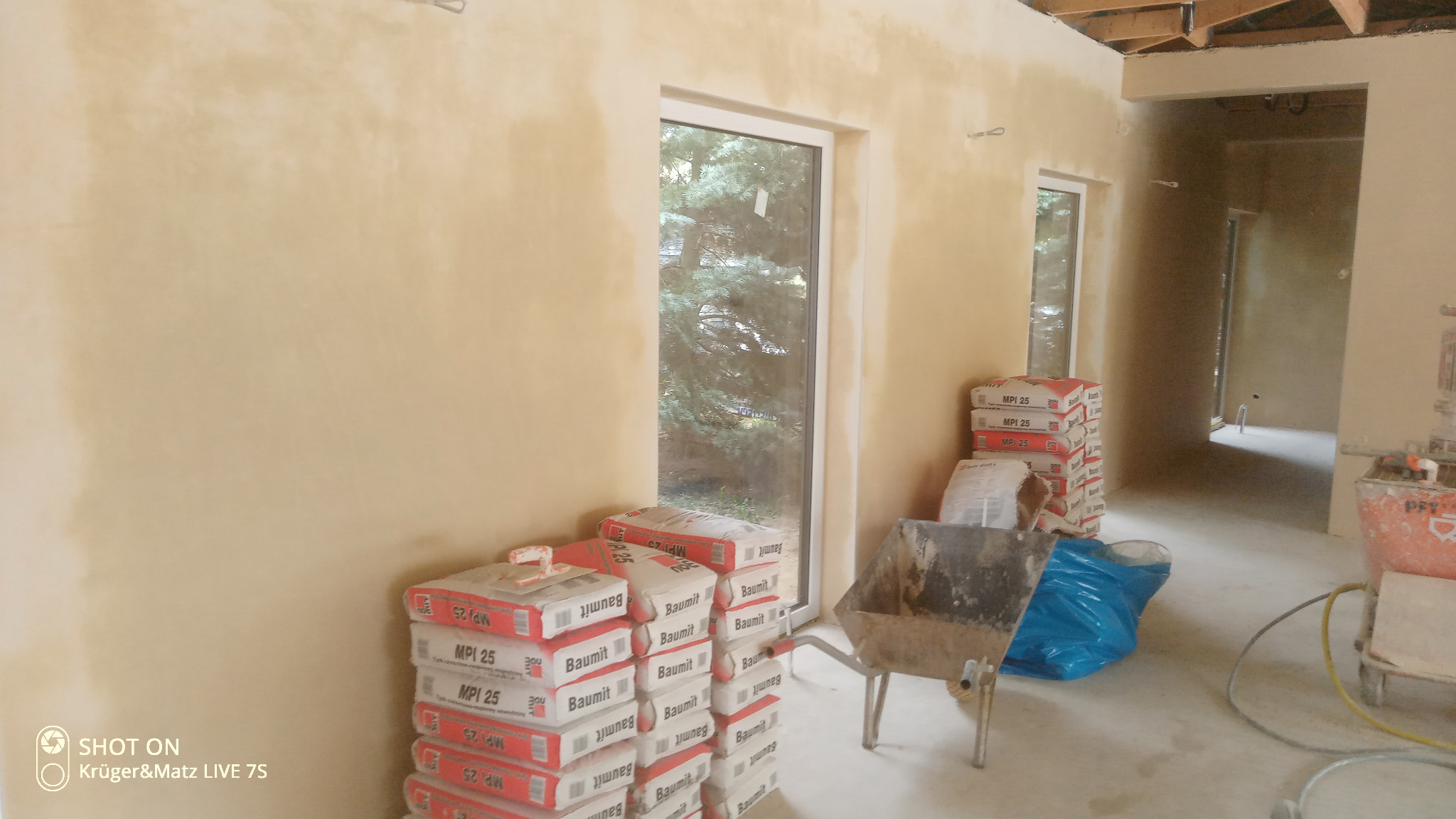 Świeżo położone jasne tynki w pomieszczeniu w Zabrzu, widoczne worki z zaprawą Baumit MPI 25, taczka i agregat tynkarski, okno z widokiem na zieleń.