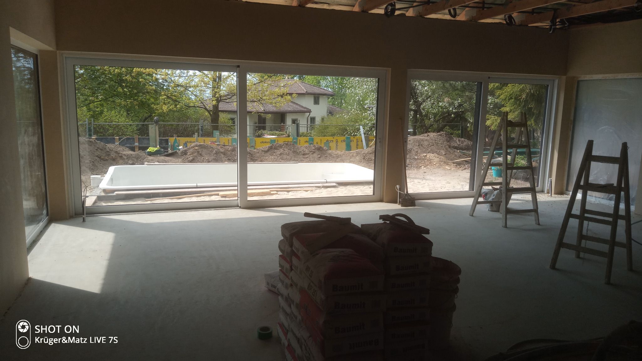 Wnętrze w trakcie tynkowania maszynowego, widoczne worki z zaprawą Baumit, drabiny malarskie oraz przez okno widok na budowę basenu w ogrodzie.