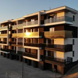 Nowoczesny budynek apartamentowy z balkonami z aluminiowymi balustradami, oświetlony promieniami zachodzącego słońca, z widocznym logo dewelopera na szczycie.