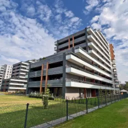 Nowoczesne apartamentowce z balkonami i szklanymi balustradami, widok z perspektywy trawnika za ogrodzeniem, błękitne niebo z chmurami w tle.