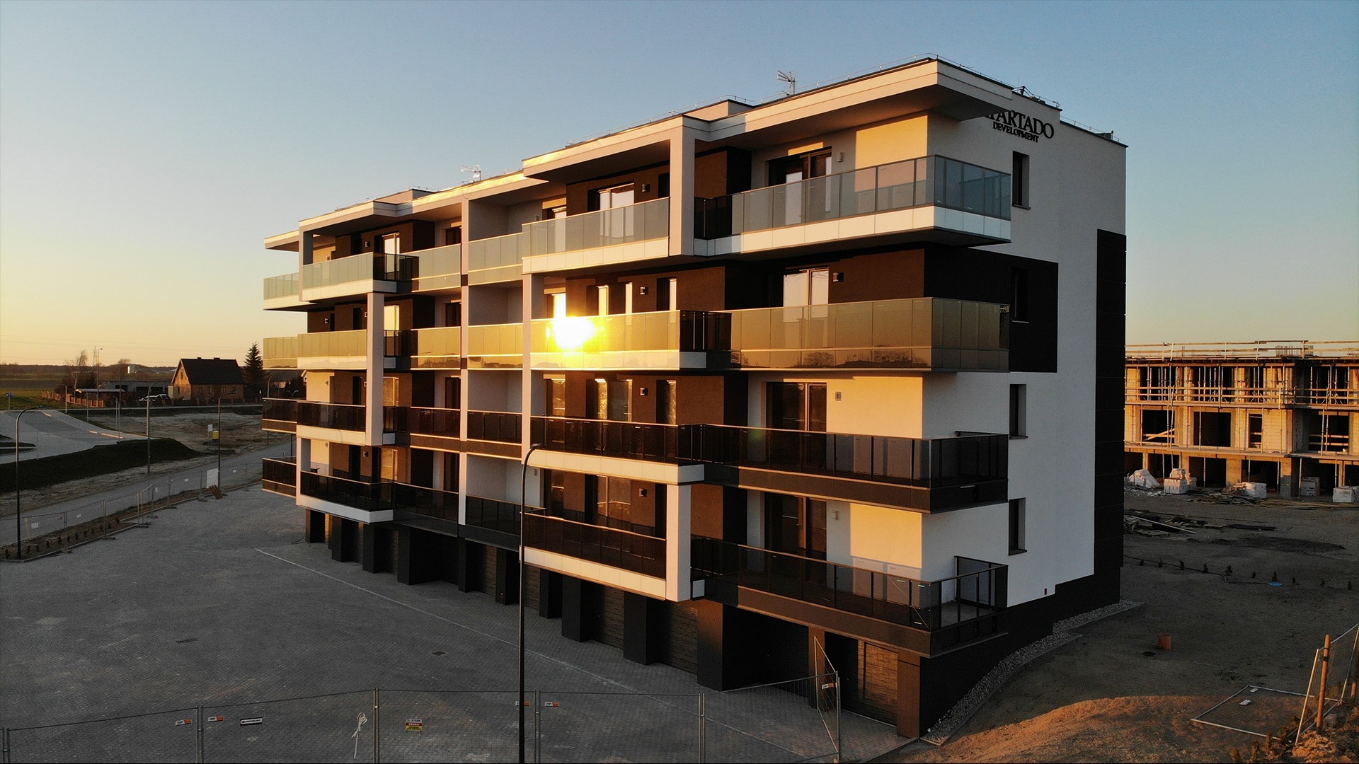 Nowoczesny budynek apartamentowy z balkonami z aluminiowymi balustradami, oświetlony promieniami zachodzącego słońca, z widocznym logo dewelopera na szczycie.