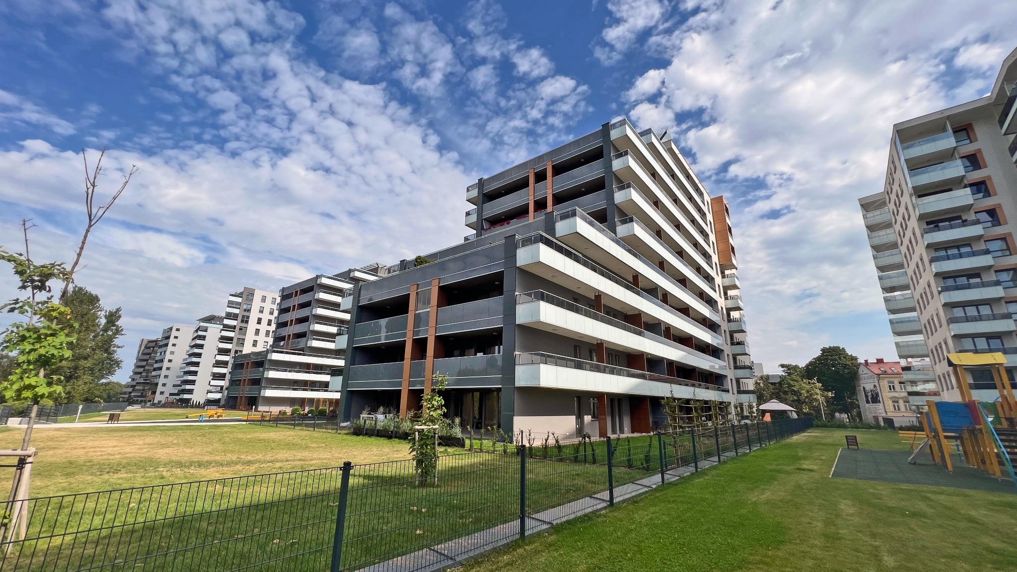 Nowoczesne apartamentowce z balkonami i szklanymi balustradami, widok z perspektywy trawnika za ogrodzeniem, błękitne niebo z chmurami w tle.
