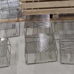 Partia metalowych, spawanych konstrukcji kratowych, prawdopodobnie stojaków lub koszy, ułożonych na betonowej posadzce obok drewnianej palety, z widocznymi oznaczeniami CFM-2 na niektórych elementach.