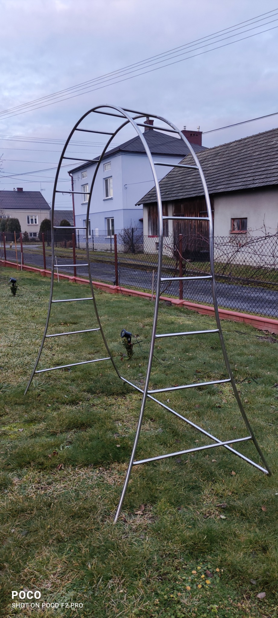 Metalowa konstrukcja łukowa z poprzeczkami na trawniku w Sędziszowie Małopolskim, w tle domy jednorodzinne i ogrodzenie z siatki.