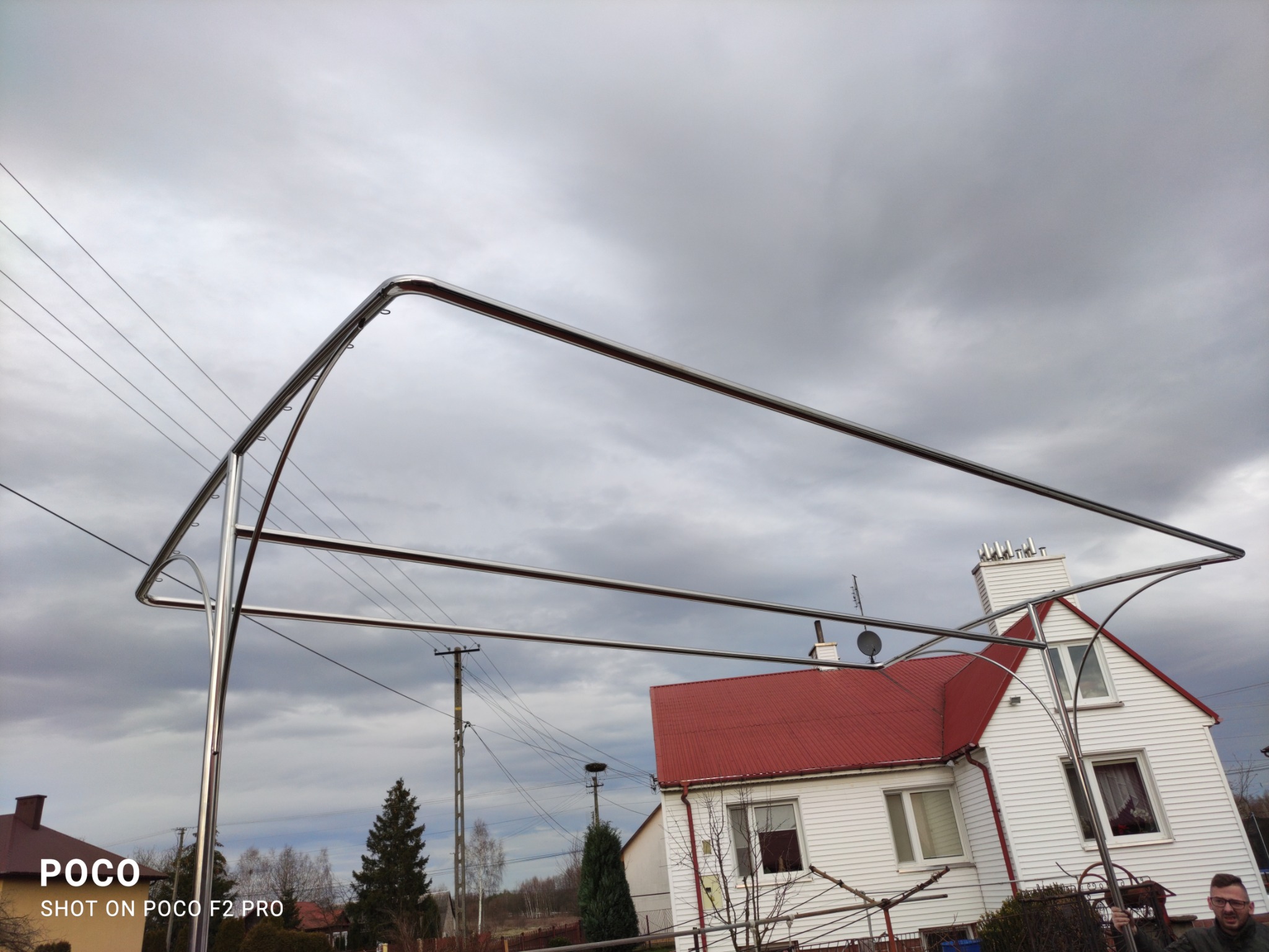Konstrukcja z polerowanej stali nierdzewnej w kształcie litery A, prawdopodobnie pod zadaszenie, na tle pochmurnego nieba i domu z czerwoną dachówką.
