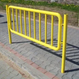 Żółta, metalowa barierka ochronna przy chodniku z kostki brukowej, mocowana na śruby.