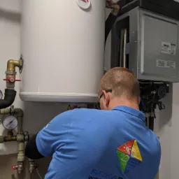 Instalator w niebieskiej koszulce z logo ROKA ENERGY, montuje biały zbiornik z manometrem i zaworami, podłączony do urządzenia grzewczego, widoczne przewody i instalacja hydrauliczna.