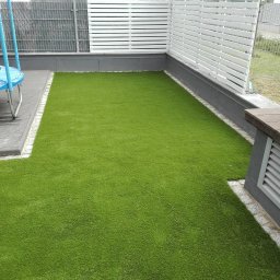 PACZO-SERVICE PAWEŁ PASZKOWSKI - Zielony trawnik z rolki otoczony szarą kostką i białym ażurowym ogrodzeniem, widok z góry. Fragment trampoliny po lewej stronie kadru.