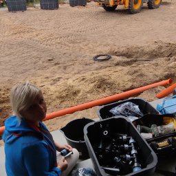 PACZO-SERVICE PAWEŁ PASZKOWSKI - Pracownik przygotowuje elementy systemu nawadniania ogrodu, w tle koparka JCB oraz ułożona kostka. Widoczne złącza, rury i narzędzia.