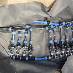 PACZO-SERVICE PAWEŁ PASZKOWSKI - Fragment instalacji nawadniającej z rurami i złączkami na czarnej folii w wykopie, widok z góry. Elementy systemu oznaczone numerami.