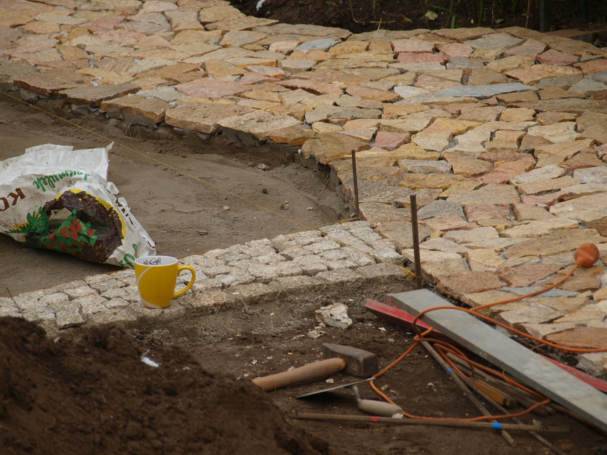 Układanie ścieżki z nieregularnych kamieni, widoczny worek z zaprawą, żółty kubek McDonald's, poziomica, młotek i inne narzędzia na ziemi.
