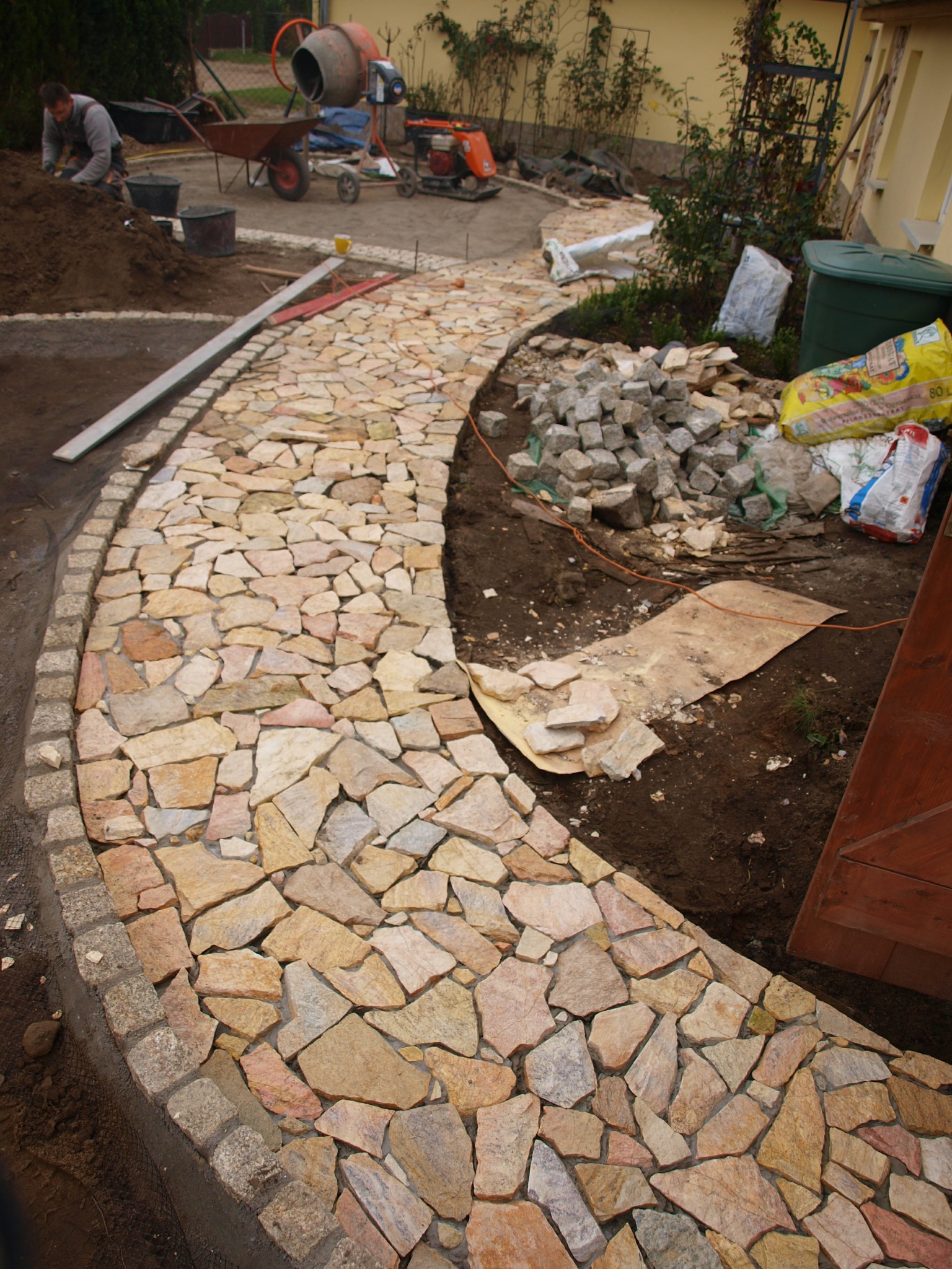 Układanie ścieżki z nieregularnych, jasnych kamieni, obrzeże z kostki granitowej, w tle pracownik, betoniarka i zagęszczarka.