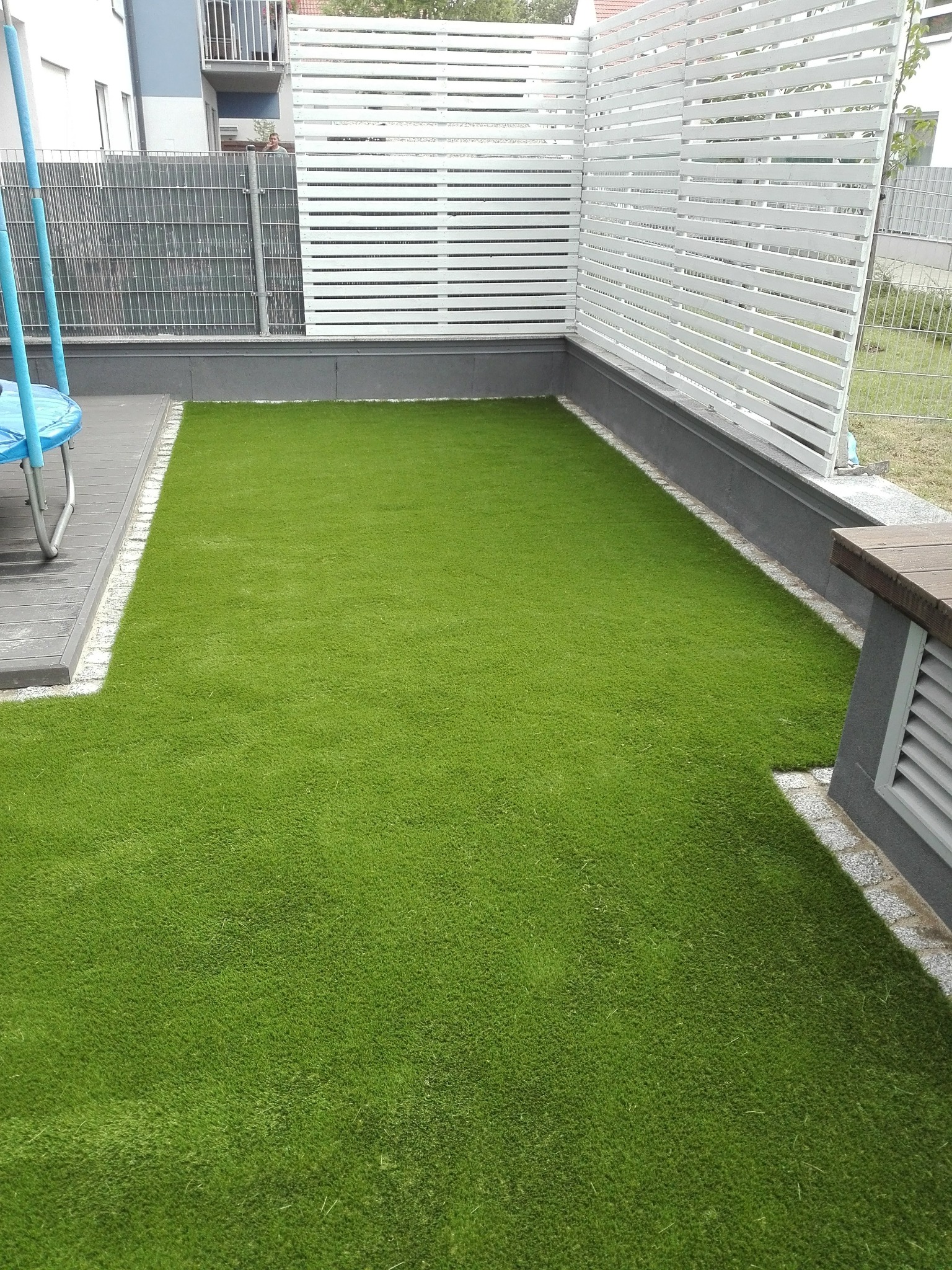 Zielony trawnik z rolki otoczony szarą kostką i białym ażurowym ogrodzeniem, widok z góry. Fragment trampoliny po lewej stronie kadru.