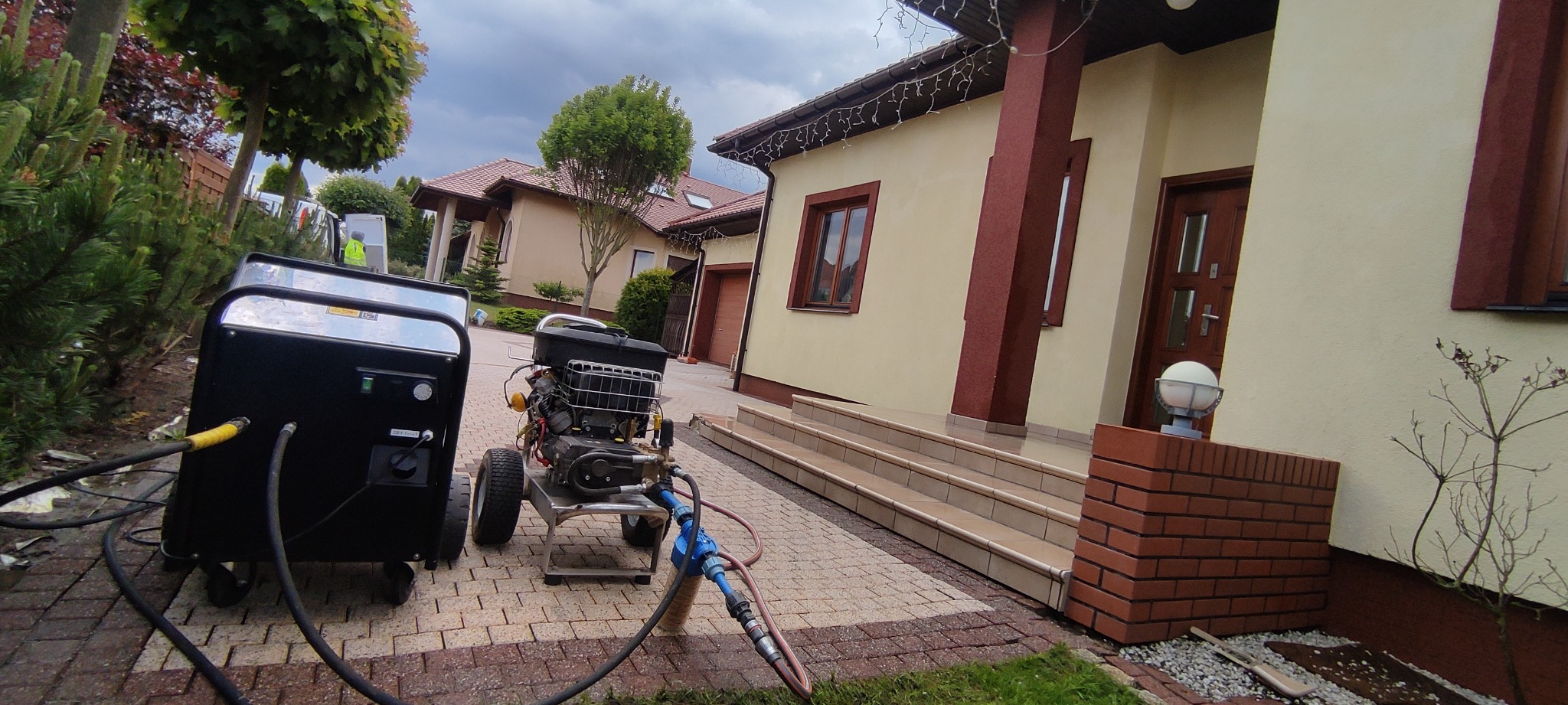 Profesjonalna myjka ciśnieniowa na kostce brukowej przed domem. Widoczne węże i elementy urządzenia. W tle dom z beżową elewacją i brązowymi akcentami.