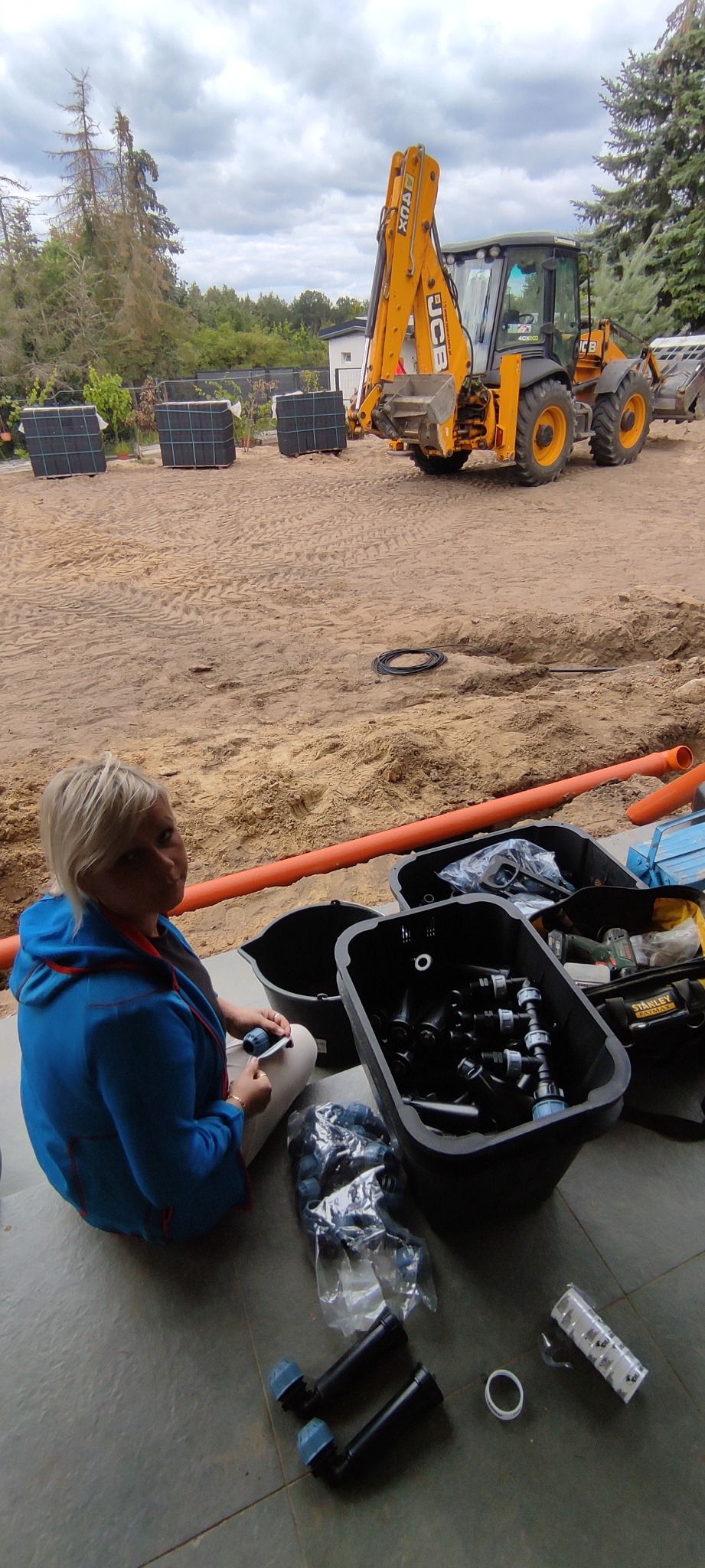 Pracownik przygotowuje elementy systemu nawadniania ogrodu, w tle koparka JCB oraz ułożona kostka. Widoczne złącza, rury i narzędzia.