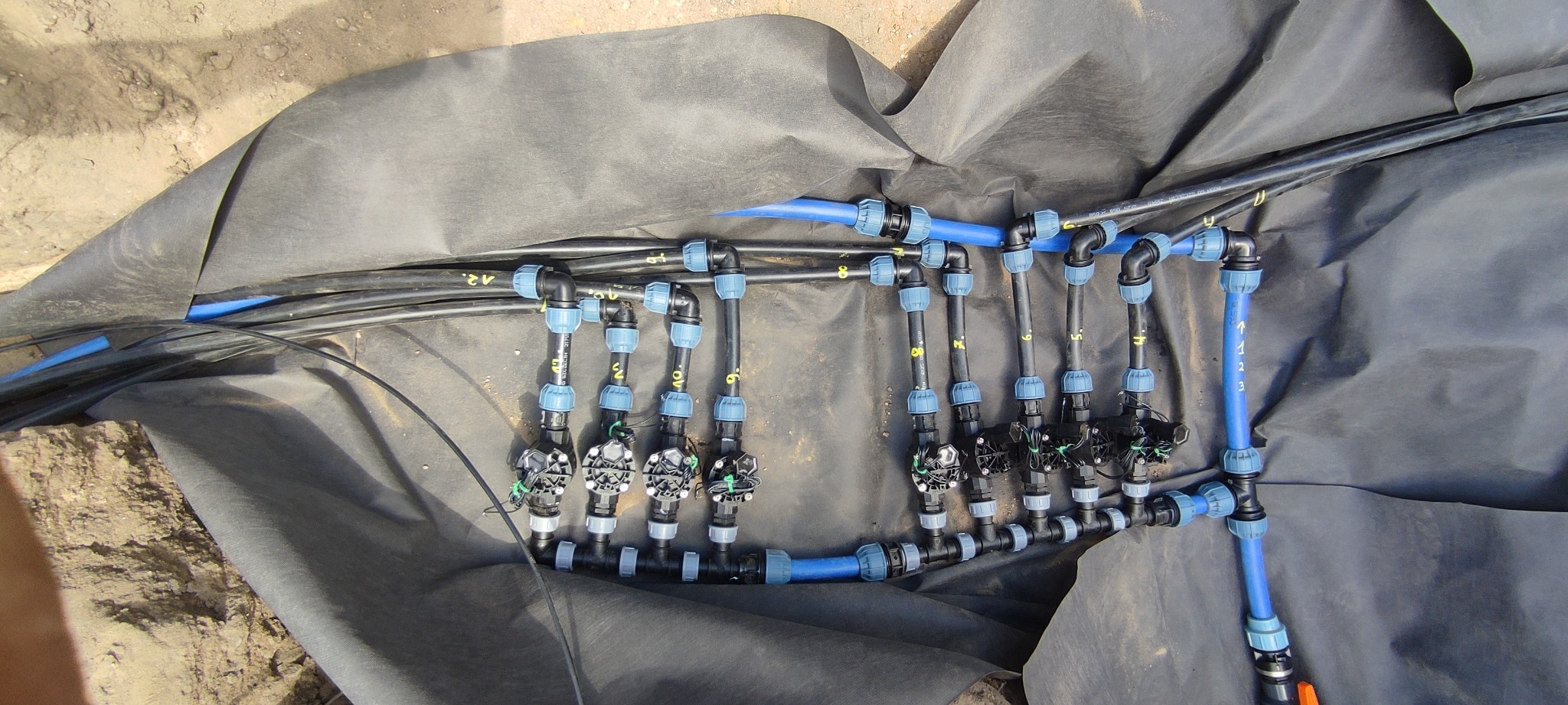 Fragment instalacji nawadniającej z rurami i złączkami na czarnej folii w wykopie, widok z góry. Elementy systemu oznaczone numerami.