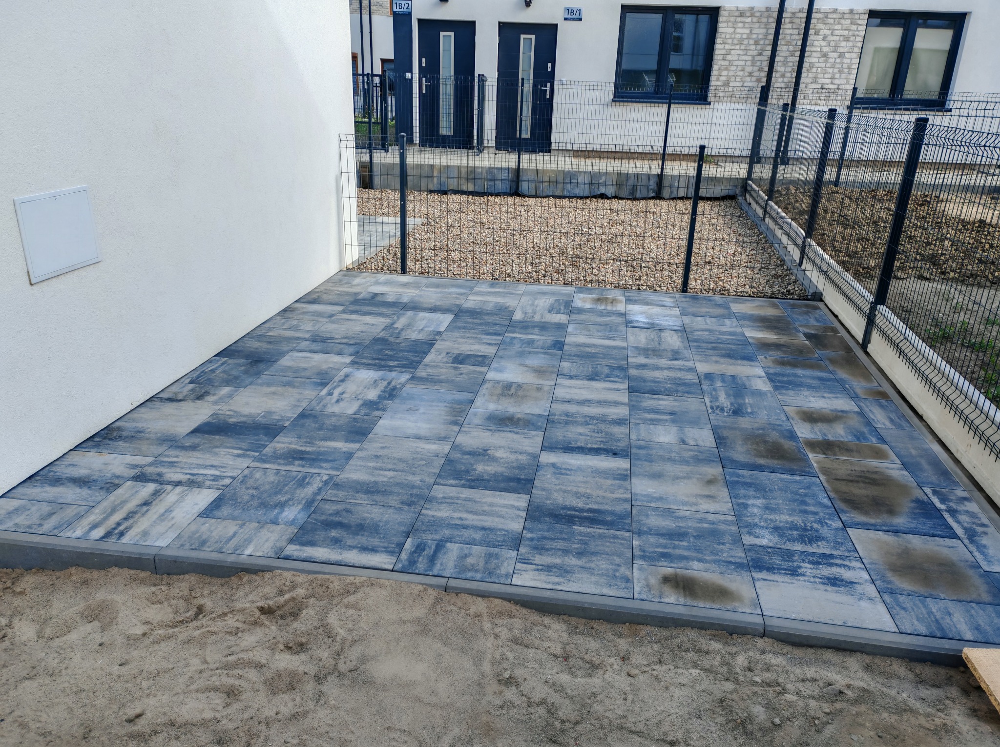 Taras wyłożony szaro-niebieską kostką brukową w geometryczny wzór, obrzeża z szarego betonu, piasek. W tle ogrodzenie z siatki i budynek z dwoma wejściami.