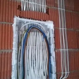 Rozdzielnica elektryczna podtynkowa z przewodami w białych peszlach zamocowanymi do ceglanej ściany, widoczne kable w kolorach niebieskim, brązowym i zielono-żółtym.