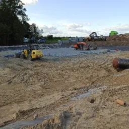 Przygotowanie terenu pod budowę: wyrównywanie gruntu, układanie geokraty i rur drenażowych, widoczny sprzęt budowlany: koparki i ładowarki.