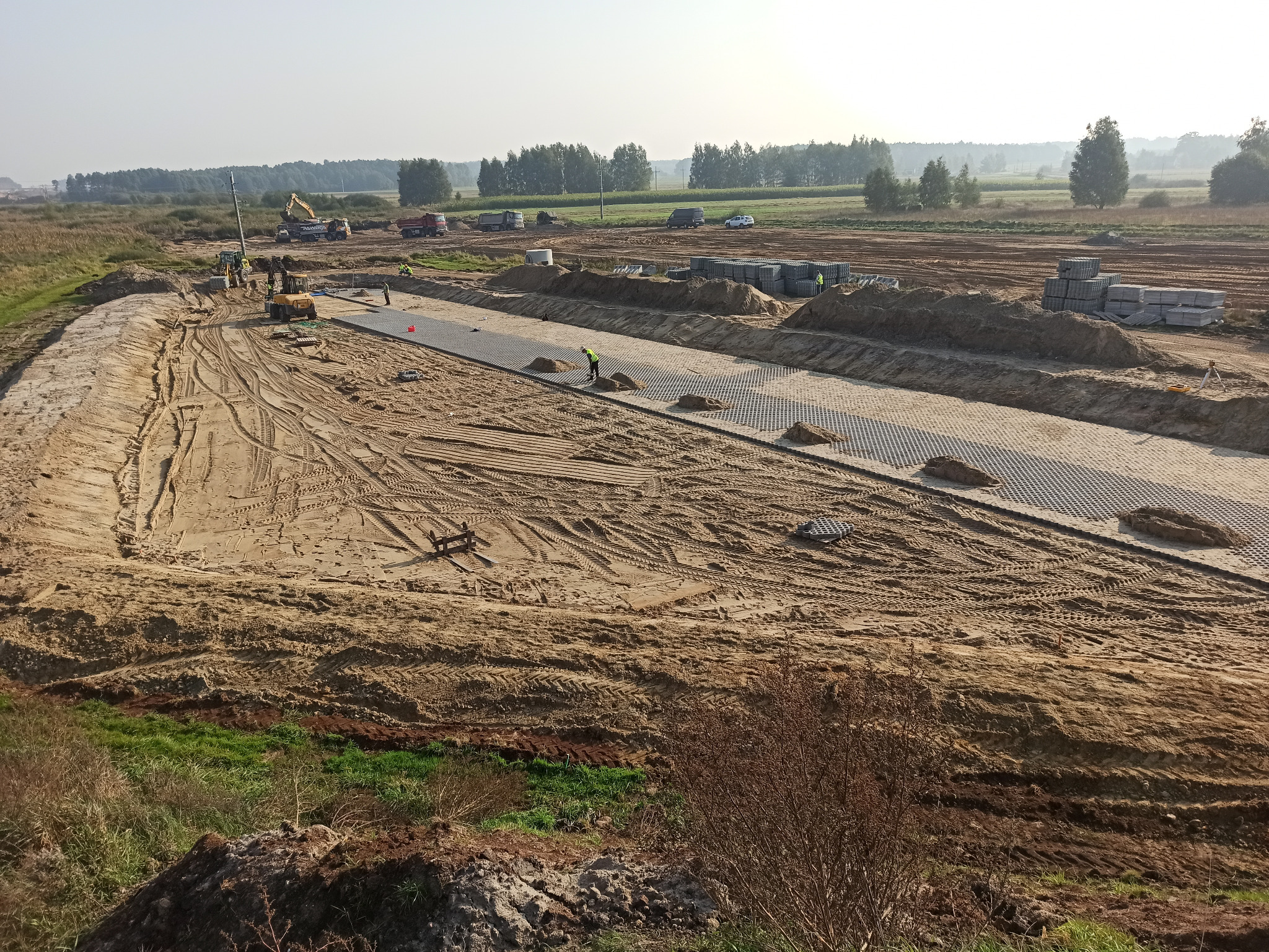Szeroki plan budowy drogi z widocznymi maszynami budowlanymi, pracownikami w kamizelkach odblaskowych, ułożoną geokratą i stertami materiałów budowlanych pod lekką mgłą.