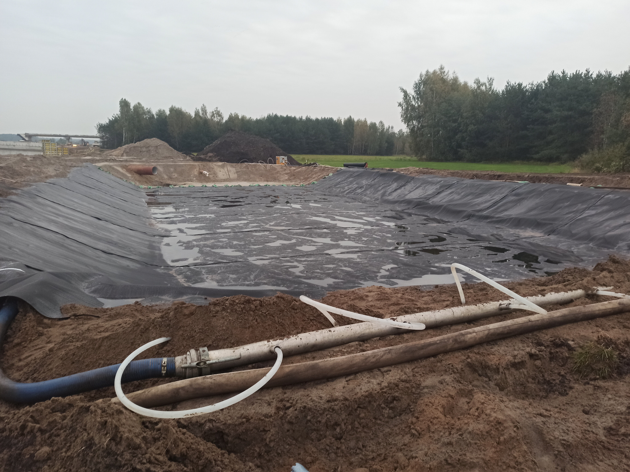 Wyłożony czarną folią wykop pod zbiornik retencyjny, wypełniony wodą, z widocznymi rurami i pompą na pierwszym planie, w tle krajobraz z drzewami i częściowo ukończoną konstrukcją.