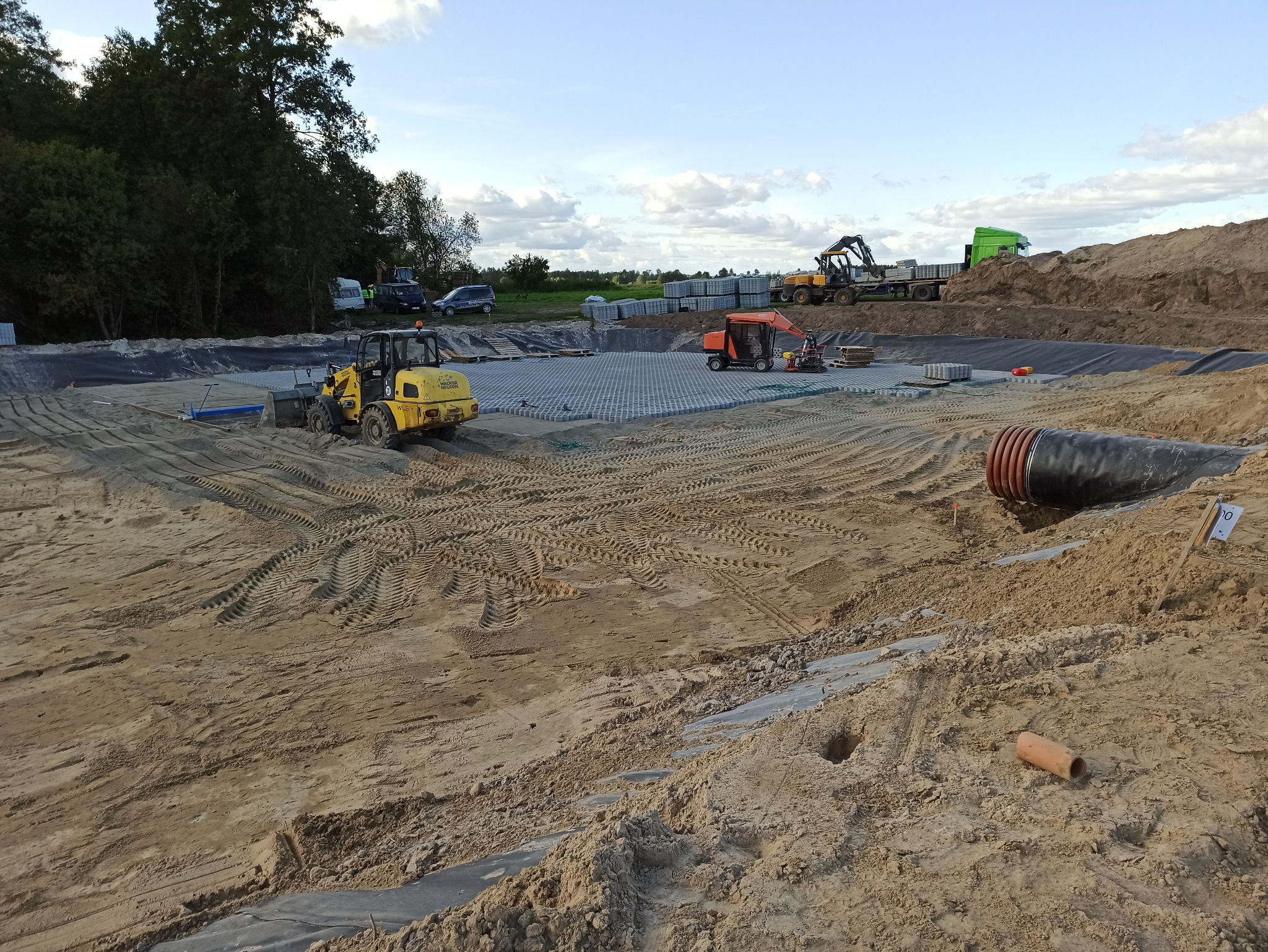 Przygotowanie terenu pod budowę: wyrównywanie gruntu, układanie geokraty i rur drenażowych, widoczny sprzęt budowlany: koparki i ładowarki.