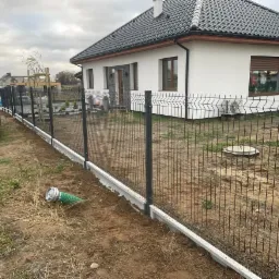 Nowoczesne ogrodzenie panelowe w kolorze grafitowym z podmur&oacute;wką betonową, otaczające dom z białej elewacji, widoczny samoch&oacute;d dostawczy i skrzynka elektryczna.