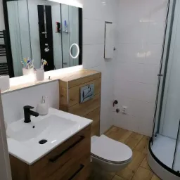 Nowoczesna łazienka z drewnopodobnymi płytkami podłogowymi i szafką pod umywalką, białe płytki ścienne, kabina prysznicowa z czarnymi ramami i podświetlane lustro okrągłe i prostokątne.